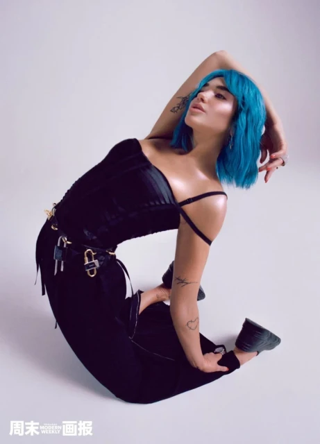 Attractive young woman with blue hair, wearing a black top and pants, poses in a kneeling position against a plain white background.