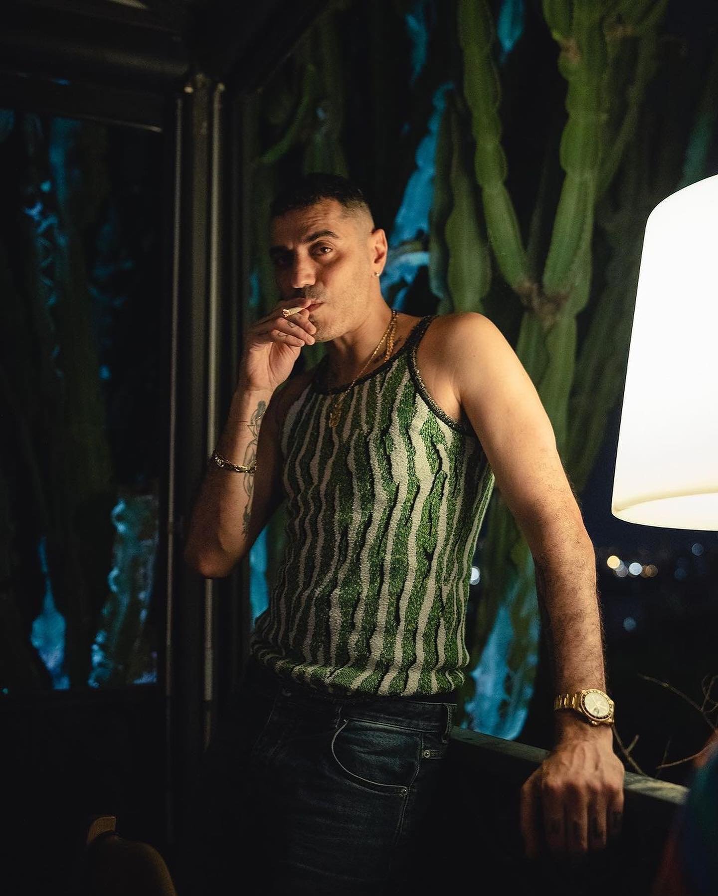 Man wearing a green striped tank top and dark jeans standing indoors with a glowing lamp and cacti in the background.