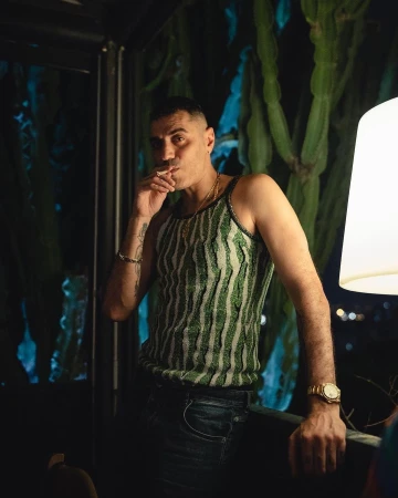 Man wearing a green striped tank top and dark jeans standing indoors with a glowing lamp and cacti in the background.
