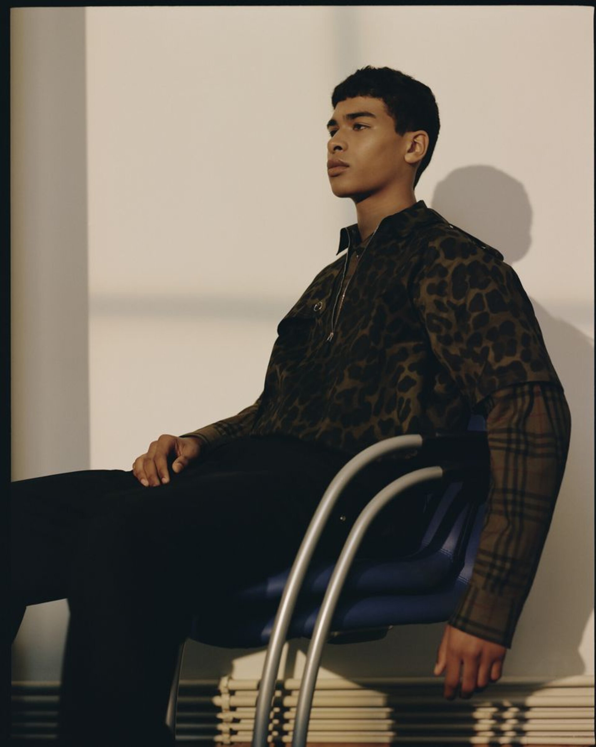 Young adult male in animal-print top and black pants seated in blue chair with silver frame against a white wall in studio setting.