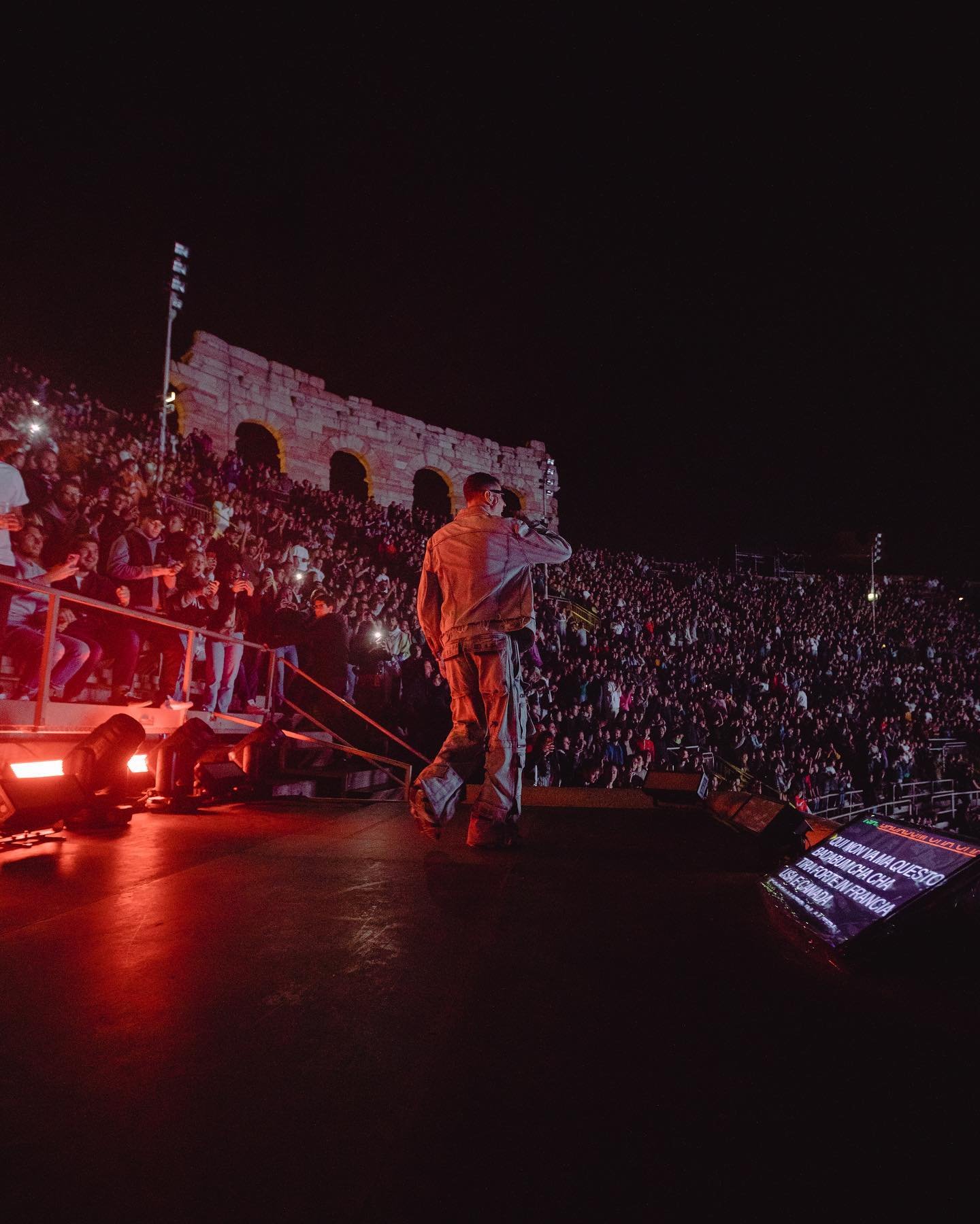 Crowd watches performer on stage at night in front of ancient architectural ruins, illuminated by red stage lights.