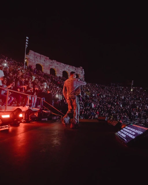 Crowd watches performer on stage at night in front of ancient architectural ruins, illuminated by red stage lights.