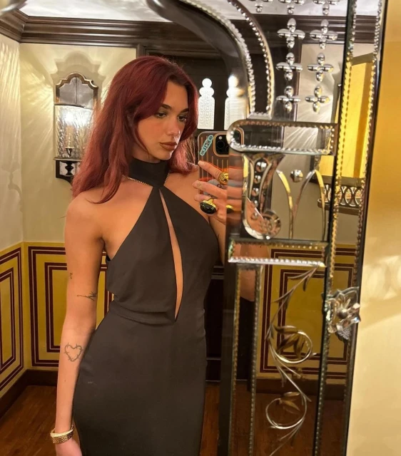 Adult woman in a black dress poses indoors, reflected in an ornate mirror with yellow-painted wall and decorative molding in the background.