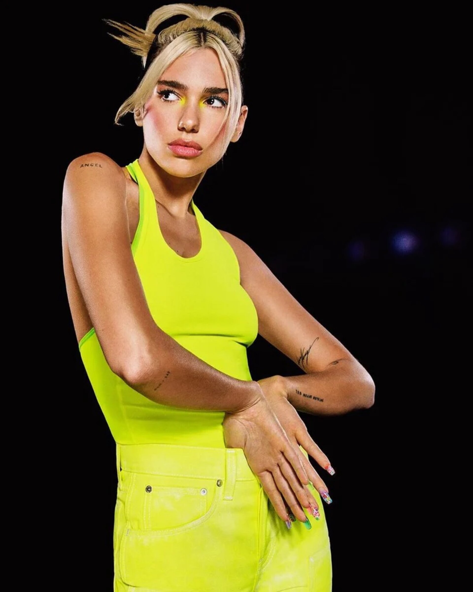 Blonde woman in neon yellow halter top and shorts poses in front of dark background with hands raised near chest level.