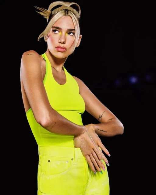 Blonde woman in neon yellow halter top and shorts poses in front of dark background with hands raised near chest level.