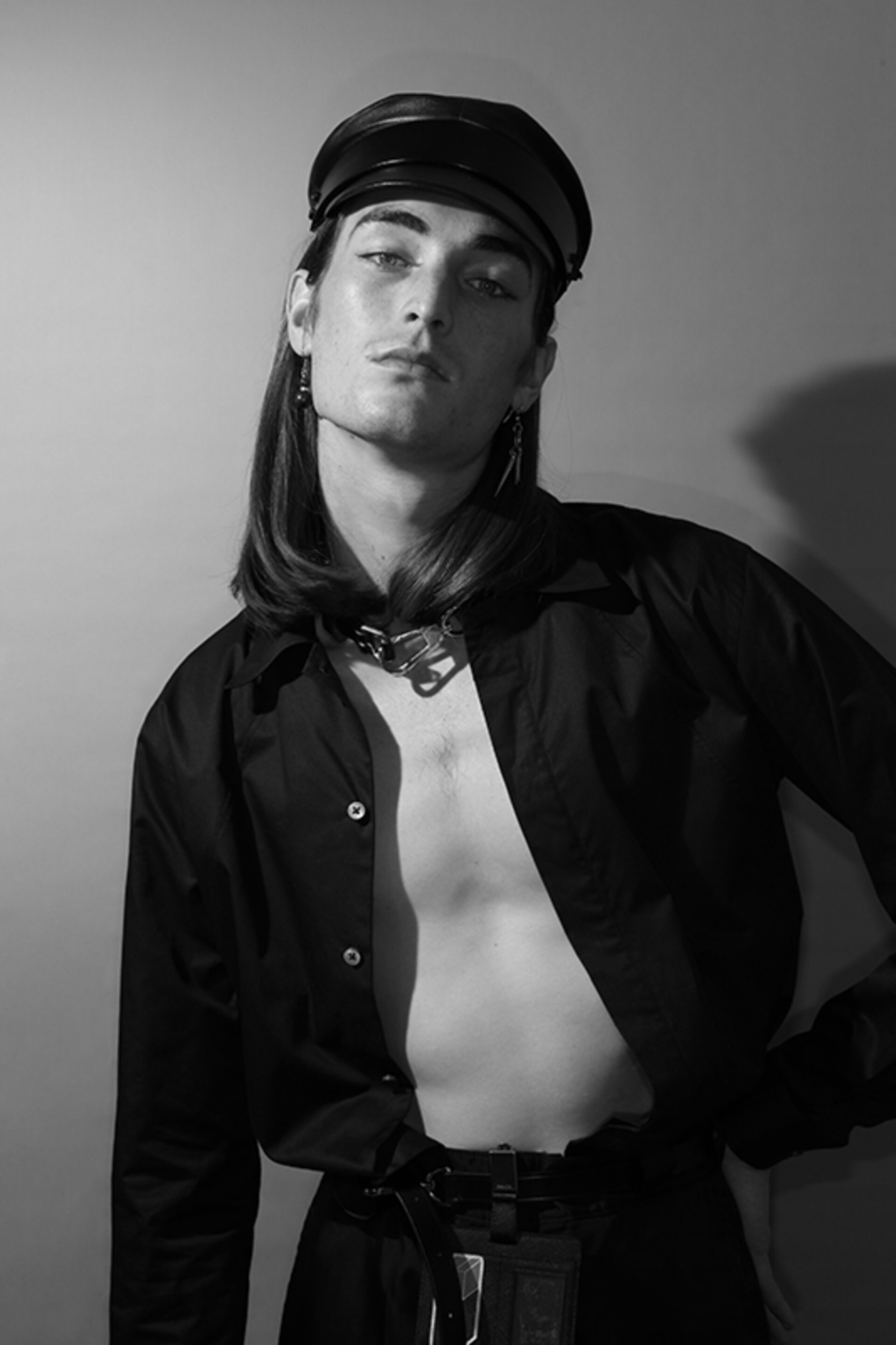 Monochrome fashion portrait of a young man in a black open shirt, silver necklace, and leather cap posing indoors against a wall.