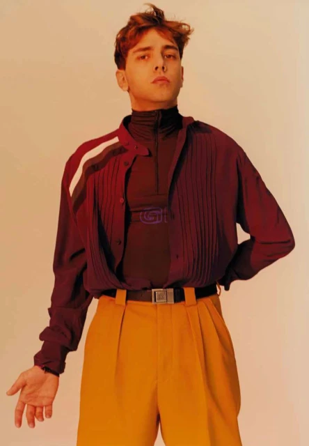 Stylish young man poses in a burgundy button-down shirt, black turtleneck, and yellow pleated trousers against an orange background.