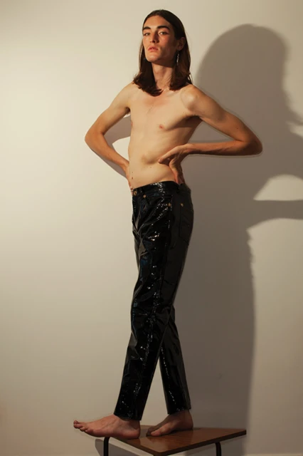 A slender, shirtless man with long brown hair stands on a wooden stool against a white wall wearing shiny black pants in a studio setting.