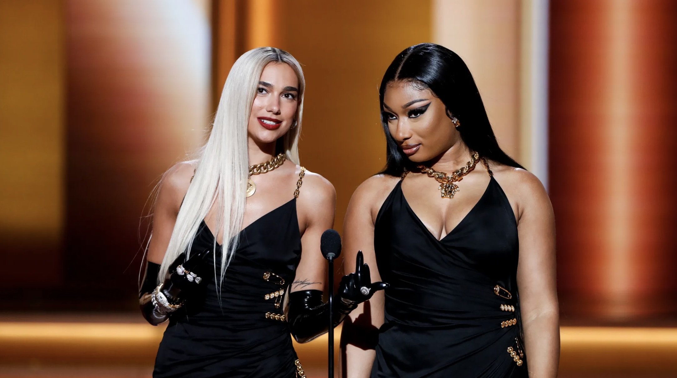 Dua Lipa and Megan Thee Stallion pose in black dresses with gold accents at an event, showcasing a blend of music and fashion.