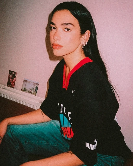 Portrait of a woman with long dark hair, wearing a black jersey and jeans, posing indoors against a light pink wall. 