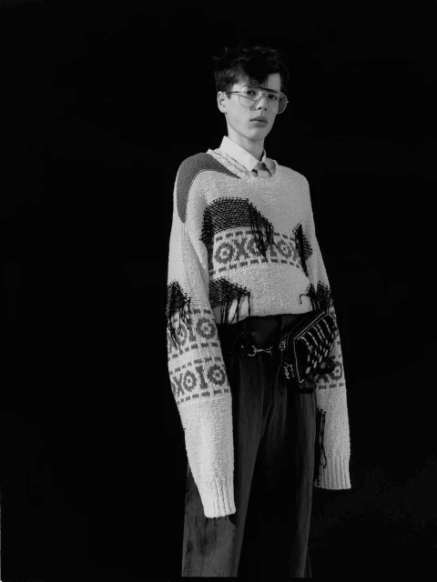 Monochrome fashion portrait of a young adult male wearing a patterned knit sweater, trousers, eyeglasses, and a small bag against a black background.