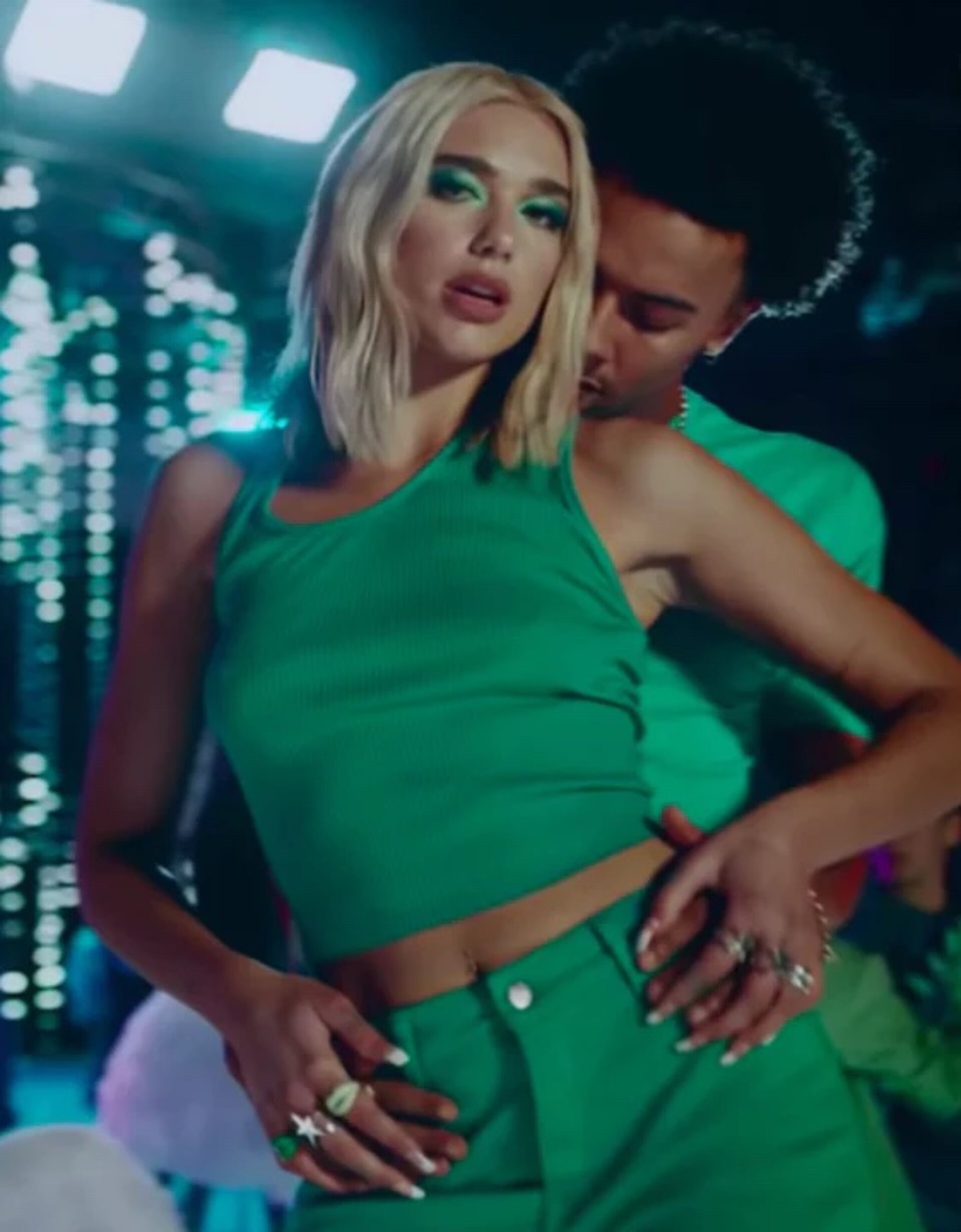 Dua Lipa in green outfit with man in green shirt, medium shot in studio with bright lighting and modern set design.