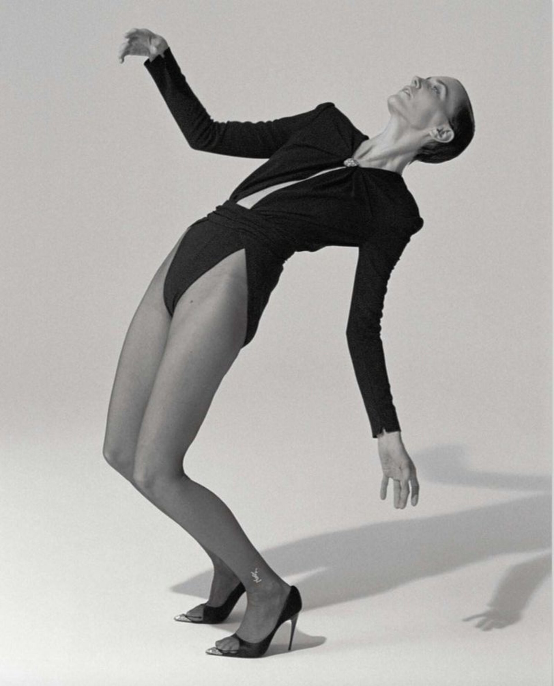 A slender adult female fashion model with a dark bodysuit and heels poses in a studio with a clean, light background.