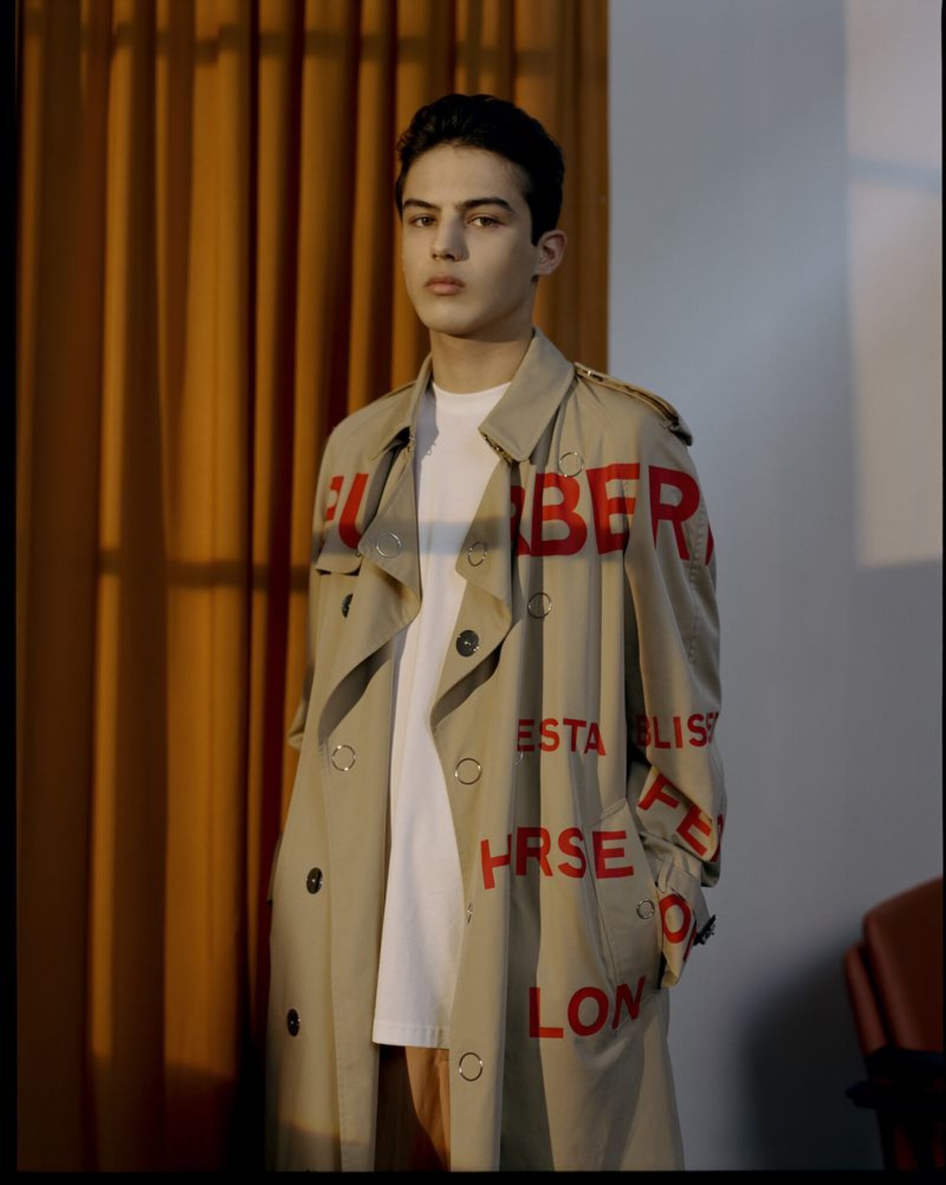Young adult posing in a beige trench coat with large red lettering, standing indoors near a beige wall and orange curtain.