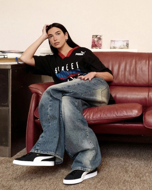 A brunette woman in a Puma shirt poses on a red couch with oversized jeans and black shoes, creating a relaxed indoor fashion shot.