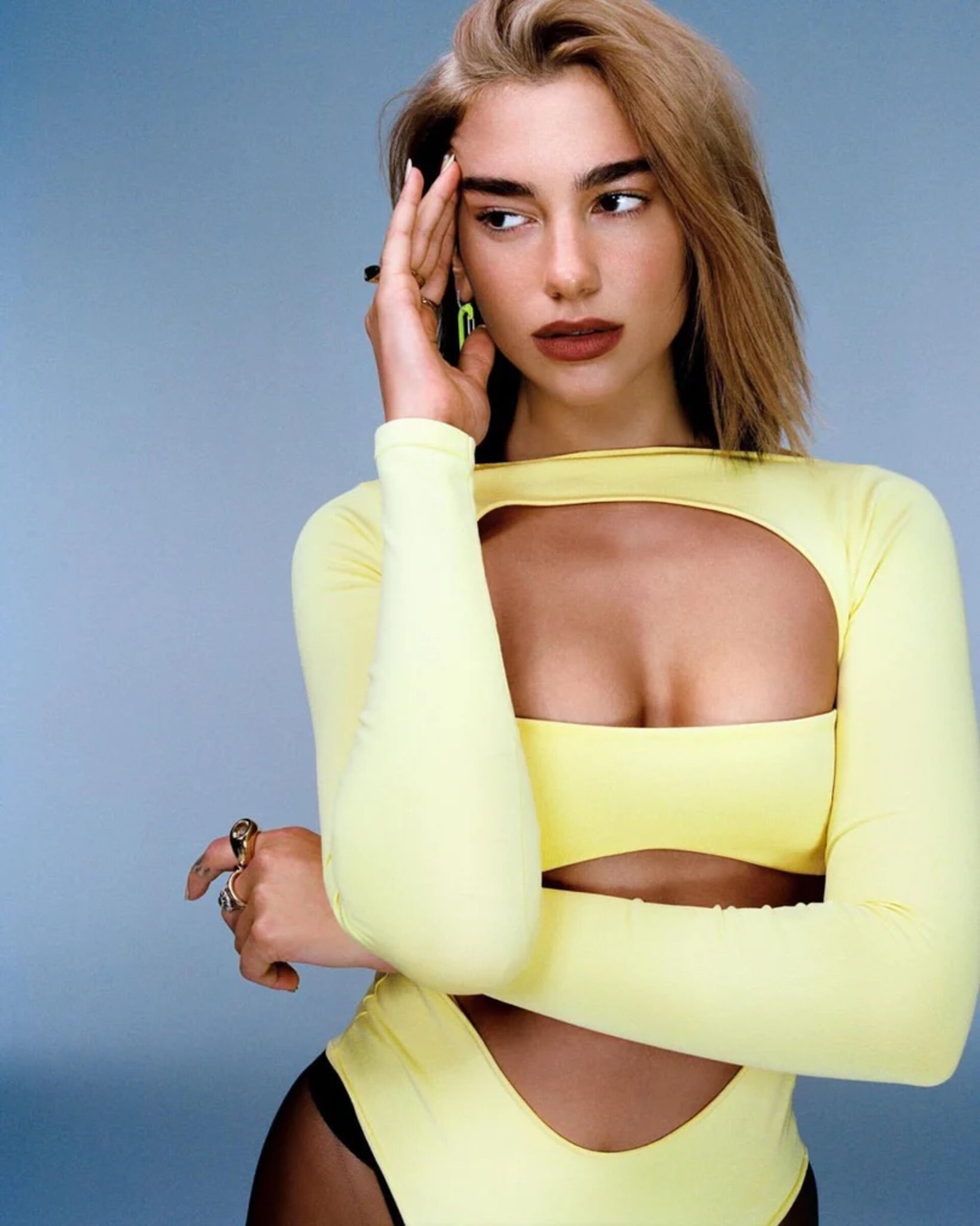 Portrait of Dua Lipa wearing a yellow cutout top and dark tights against a plain grey background