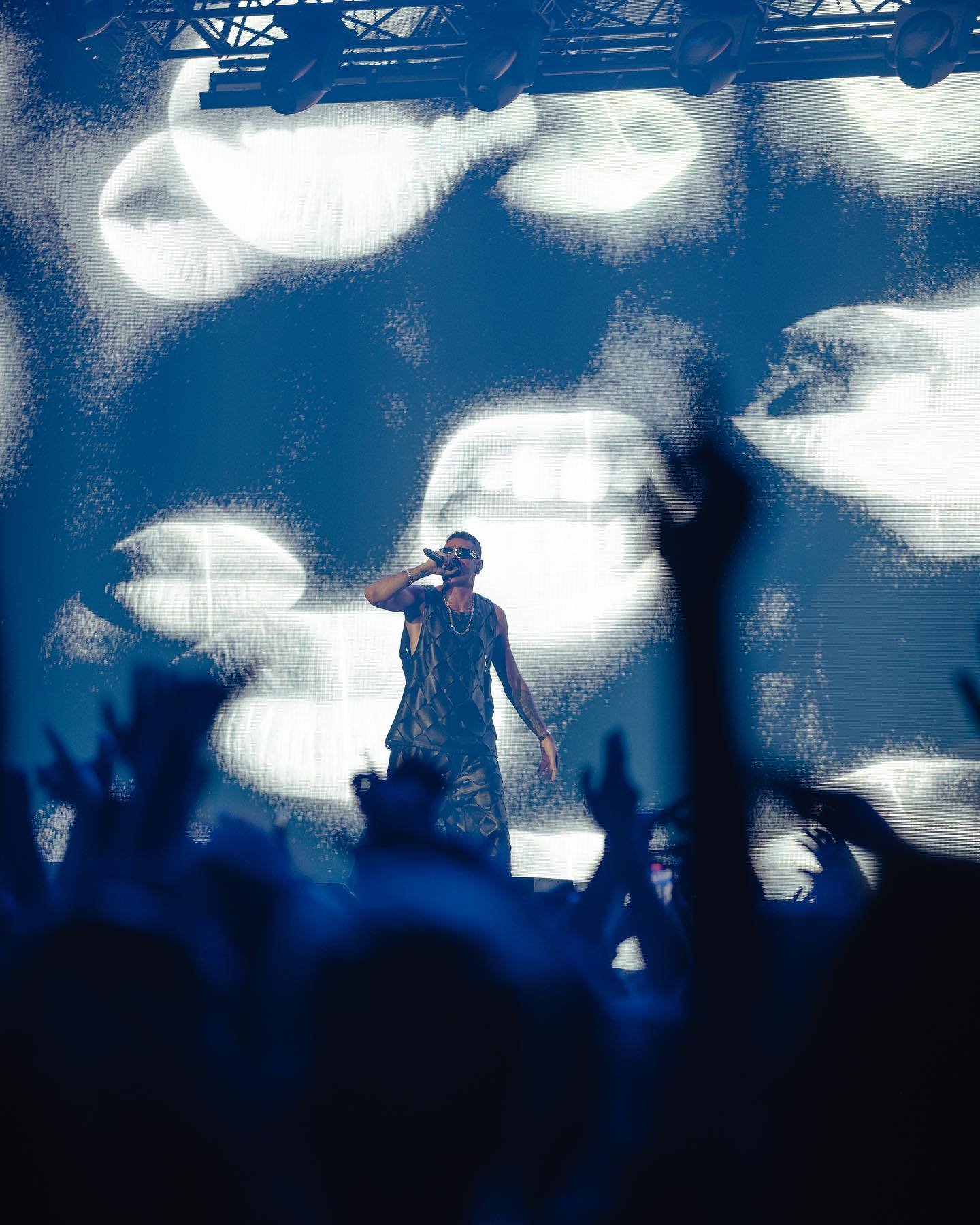 Man sings on stage in a sleeveless shirt at a concert with a background screen and silhouetted crowd, bathed in bright blue light.