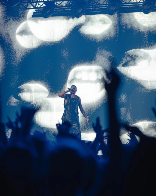 Man sings on stage in a sleeveless shirt at a concert with a background screen and silhouetted crowd, bathed in bright blue light.