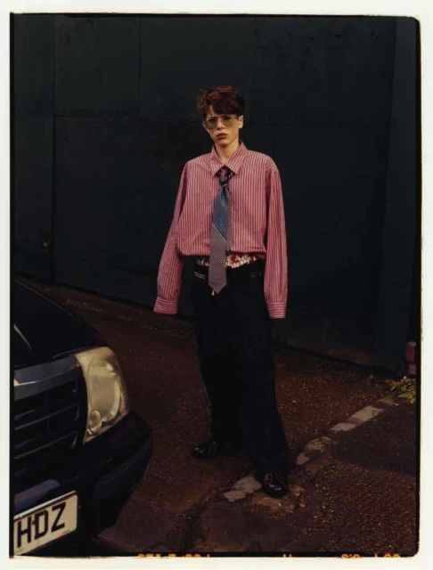 Stylish adult poses in a red-striped shirt and dark pants in an urban setting with a car parked on a city street.