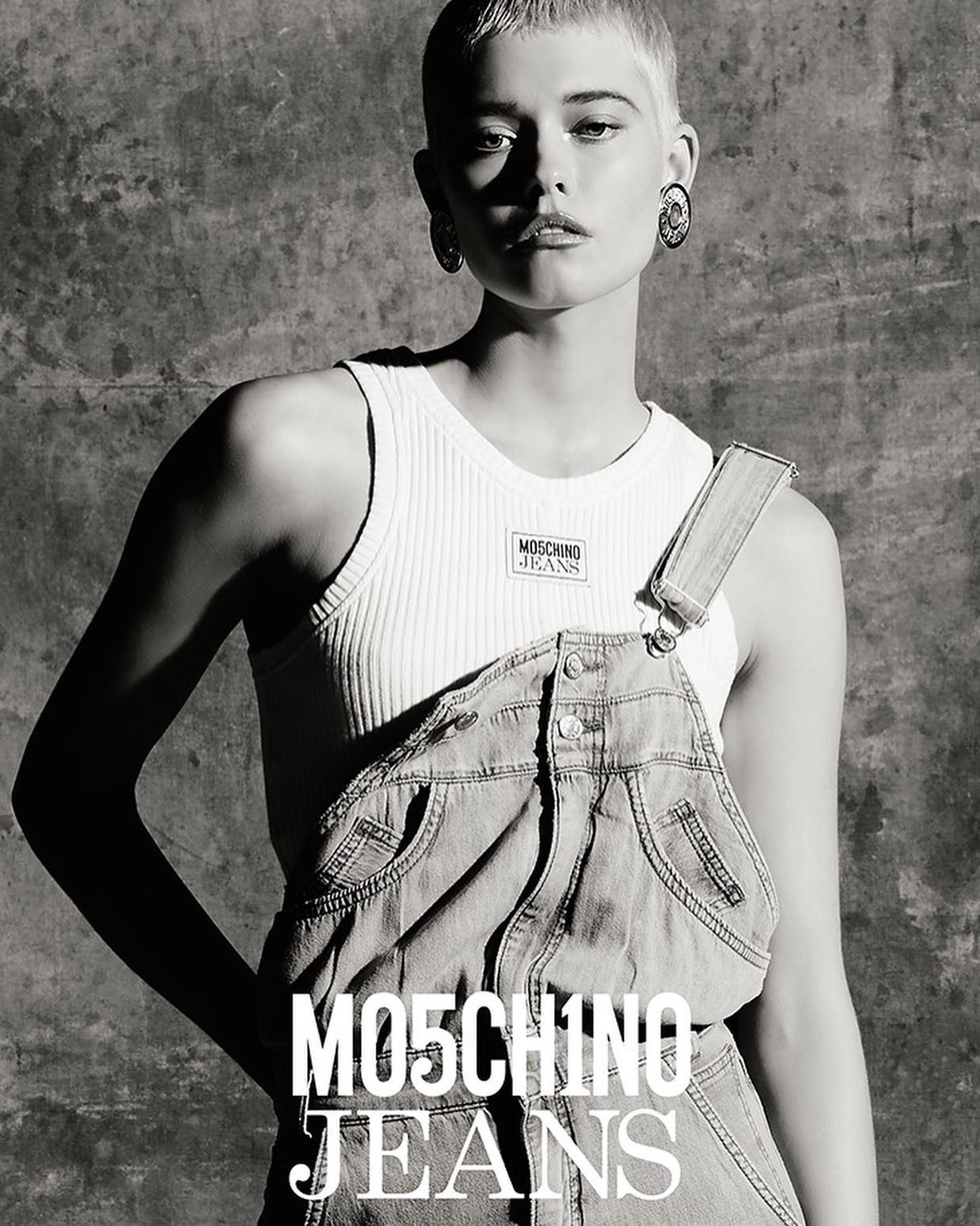 Black and white portrait of a model with short blonde hair wearing denim overalls and a ribbed tank top