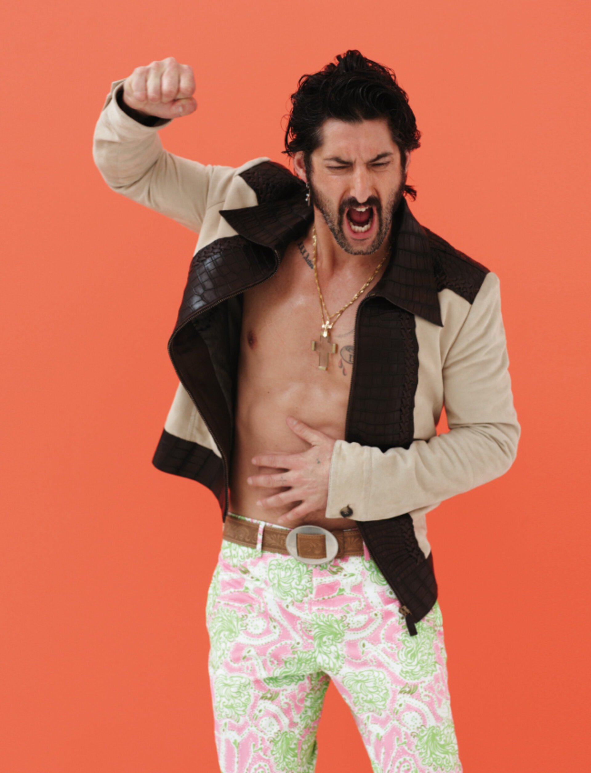 Stylish man with an open jacket, patterned pants, and a gold chain raises his fist against an orange backdrop in a studio setting.