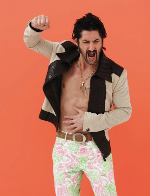 Stylish man with an open jacket, patterned pants, and a gold chain raises his fist against an orange backdrop in a studio setting.