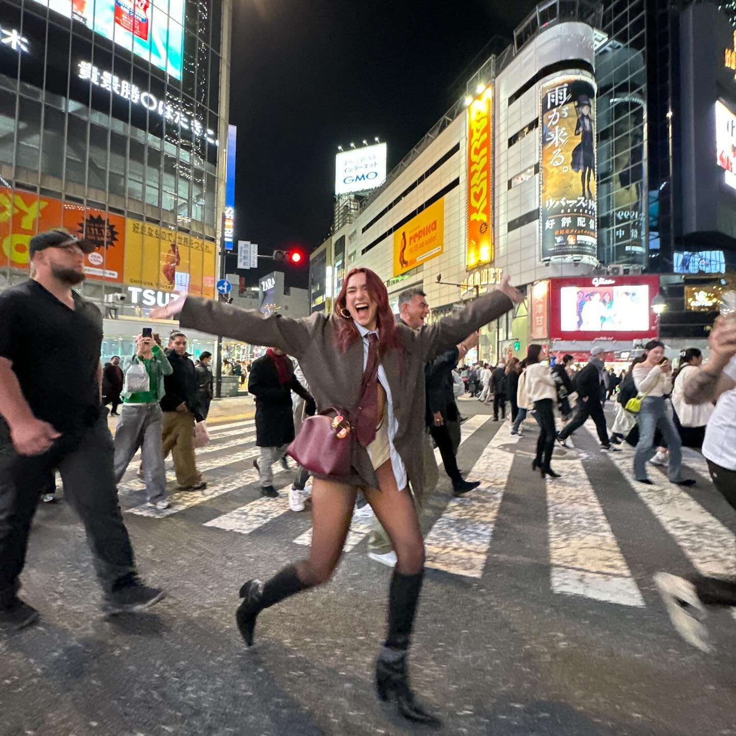 Woman in a brown suit jubilantly runs across a Tokyo street at night, surrounded by crowds, vibrant lights, and large video billboards.