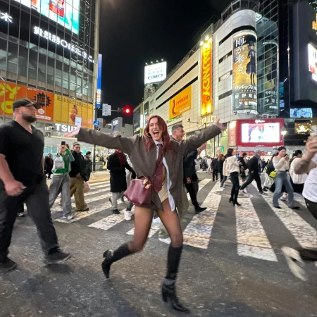 Woman in a brown suit jubilantly runs across a Tokyo street at night, surrounded by crowds, vibrant lights, and large video billboards.