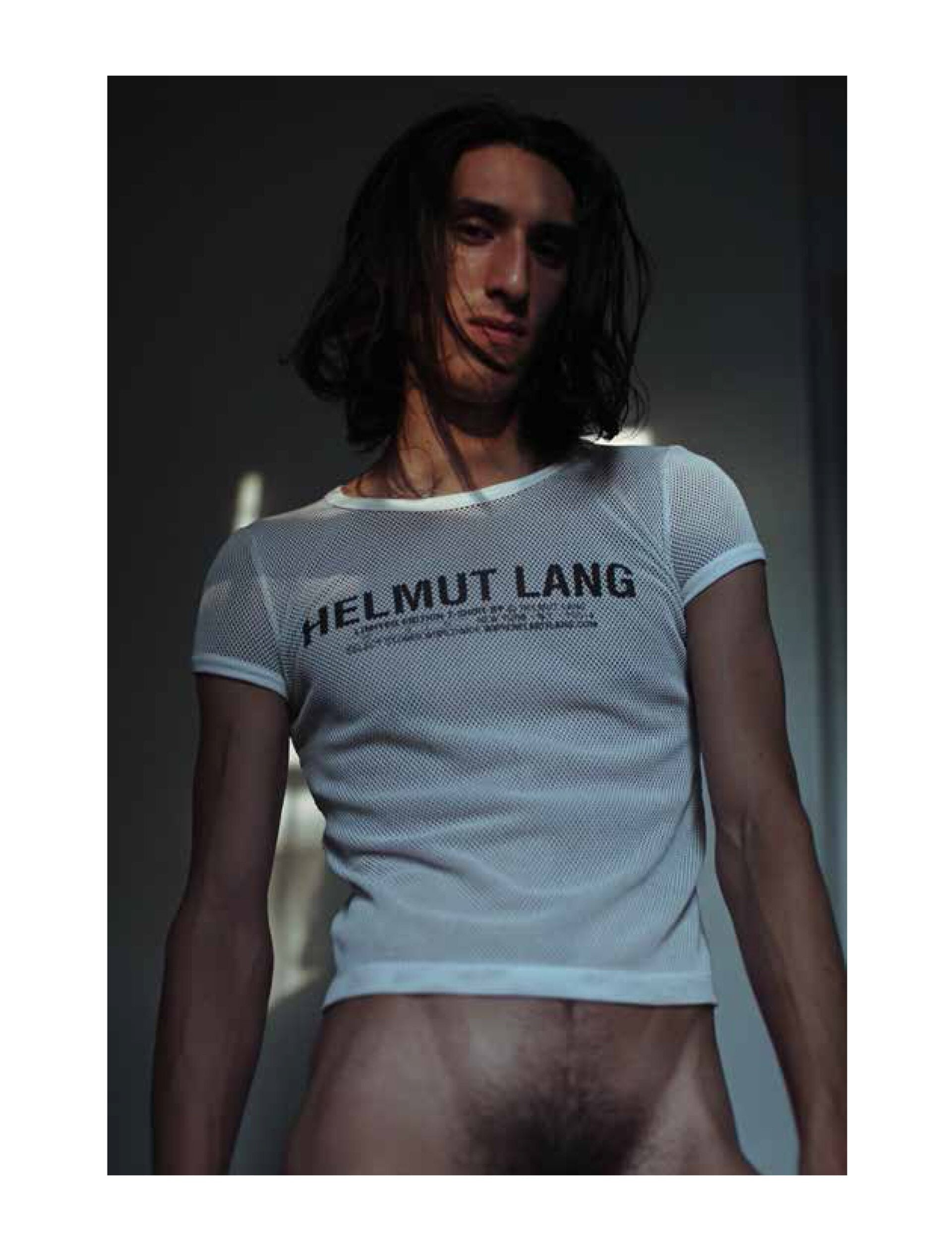 Portrait of a male model with dark hair, wearing a white mesh HELMUT LANG shirt in an indoor setting with natural lighting.