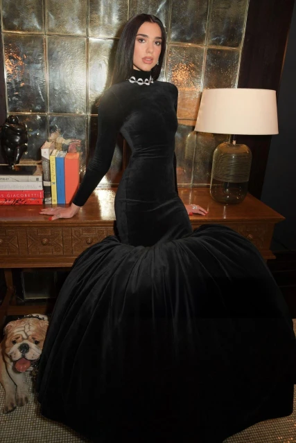 Dua Lipa poses in a long black velvet dress with a full skirt, leaning on a wooden desk in an indoor setting with neutral colors.