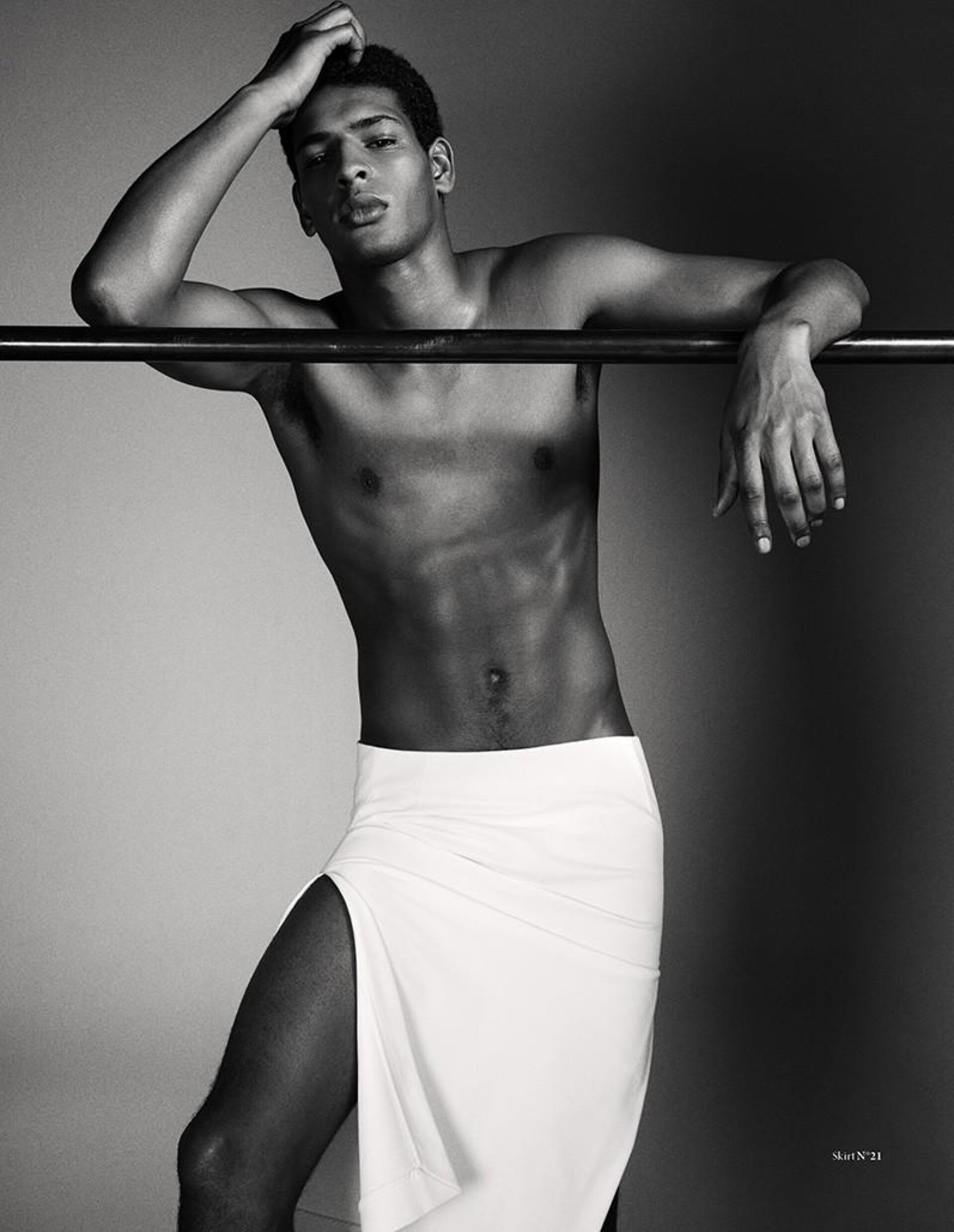 Striking black and white portrait of a shirtless, muscular Black man in a long white skirt, leaning against a metal bar, artistic fashion.
