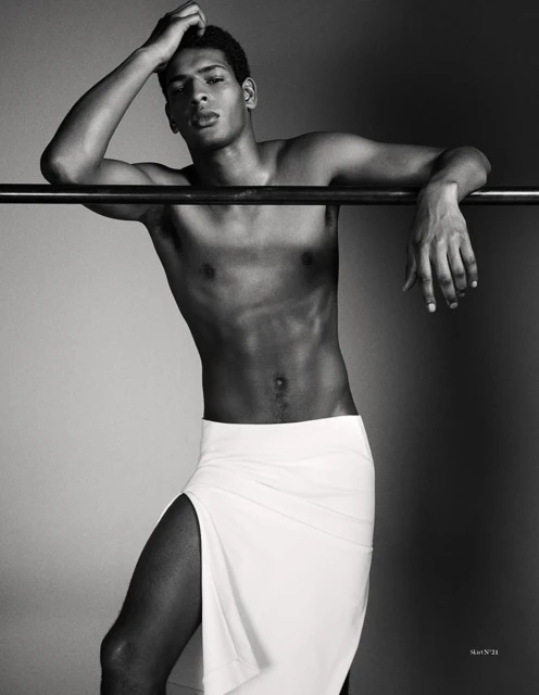 Striking black and white portrait of a shirtless, muscular Black man in a long white skirt, leaning against a metal bar, artistic fashion.