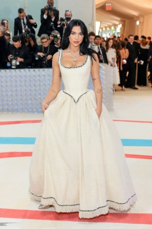 Dua Lipa at a gala event wearing a large, formal white dress with black trim. She is wearing jewelry, and has dark hair.