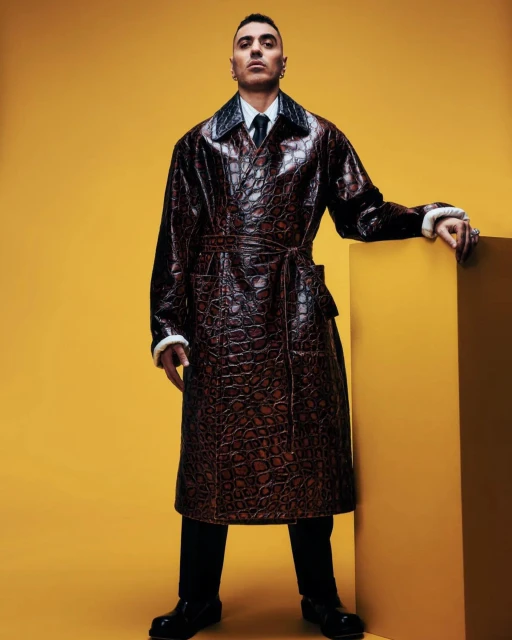 Fashionable adult male wearing a brown reptile-patterned coat in a studio with a yellow background, complemented by black pants and shoes.