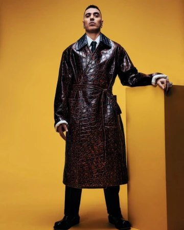 Fashionable adult male wearing a brown reptile-patterned coat in a studio with a yellow background, complemented by black pants and shoes.