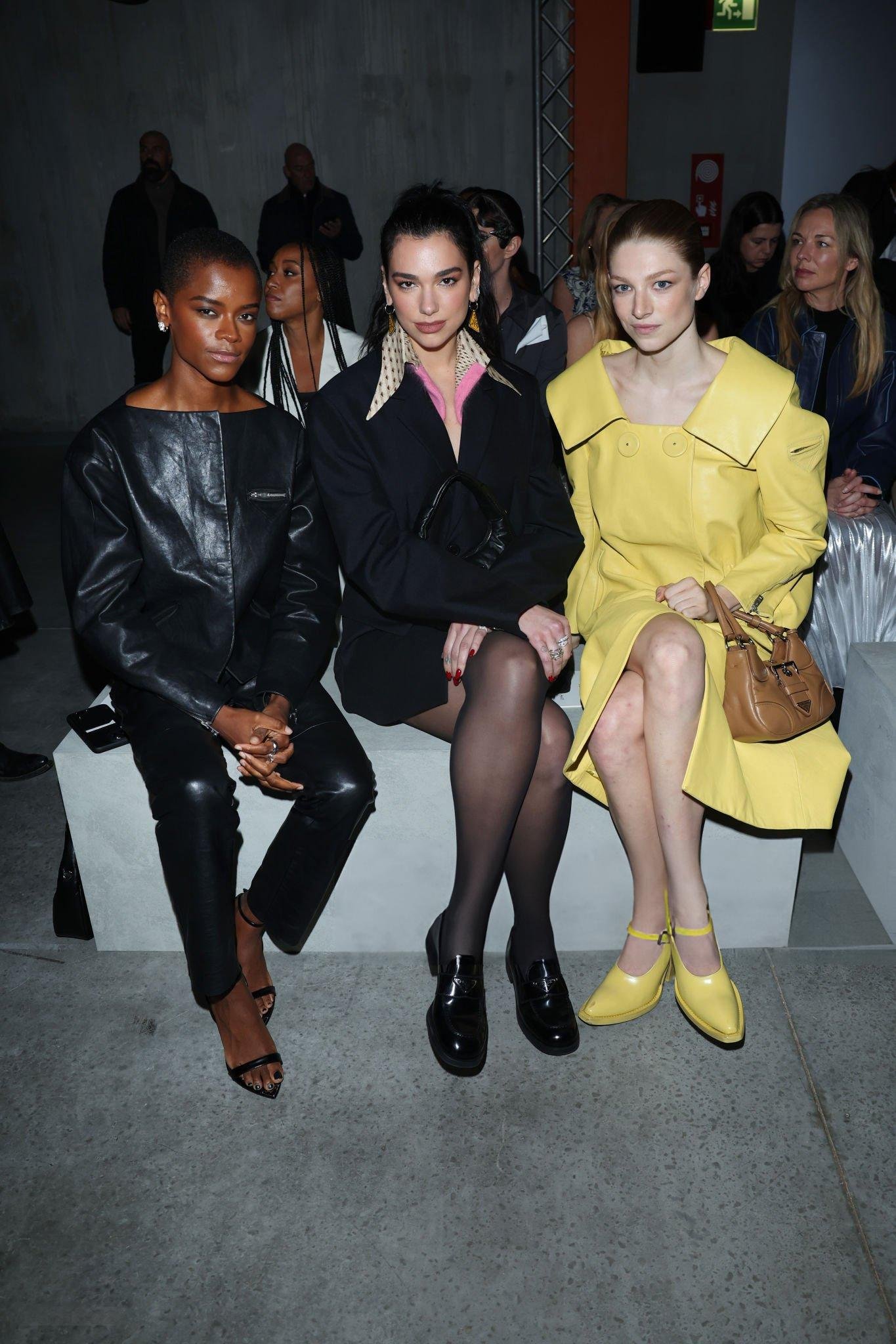 Three women, Letitia Wright, Dua Lipa, and Sadie Sink, sit in the front row at a fashion show, showcasing unique outfits and individual style.