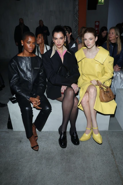 Three women, Letitia Wright, Dua Lipa, and Sadie Sink, sit in the front row at a fashion show, showcasing unique outfits and individual style.