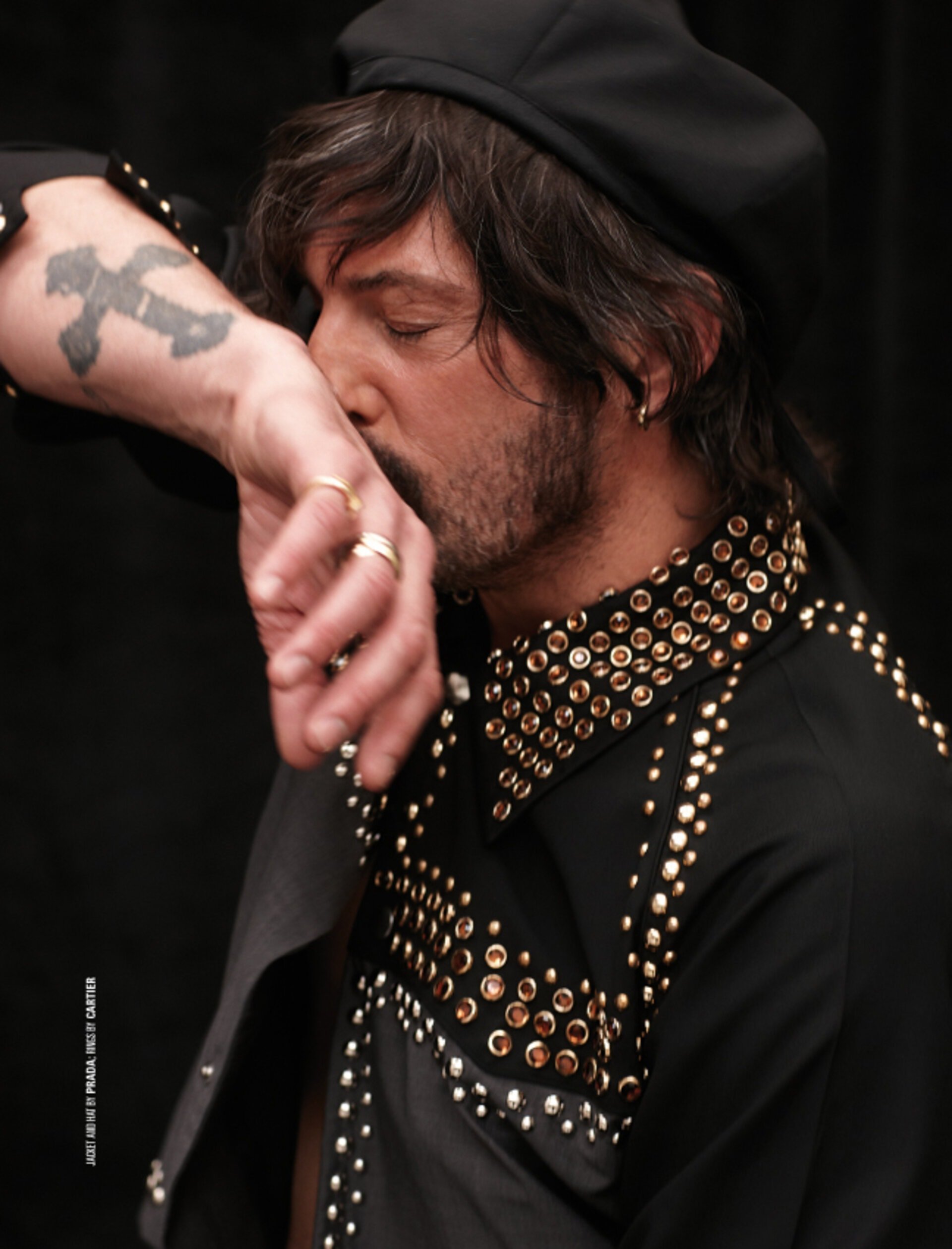 Close-up of a man wearing a black shirt with studded embellishments and a black beret, kissing the inside of his forearm with a tattoo.