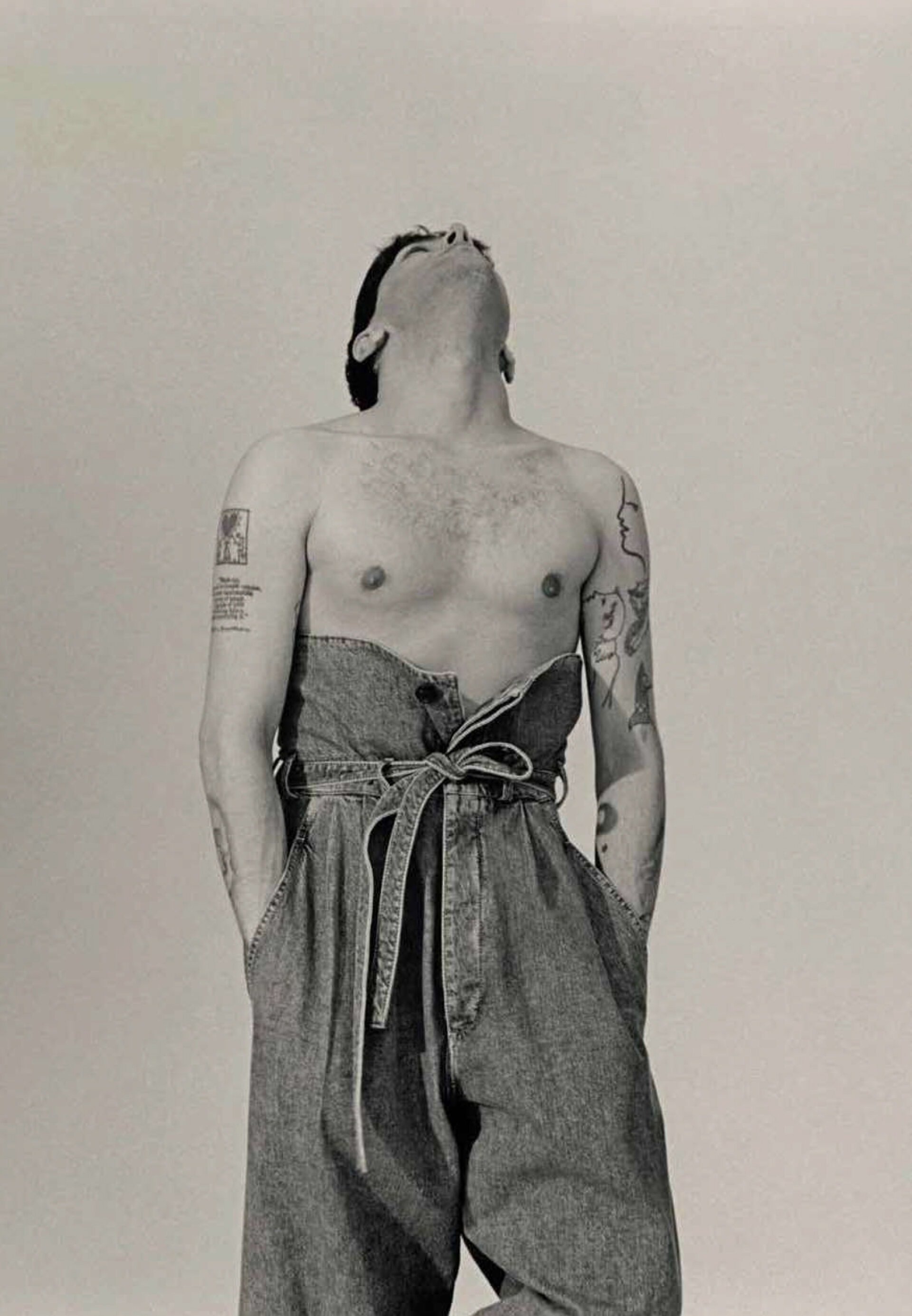 Monochrome portrait of a topless adult male with tattoos, wearing denim overalls, looking upwards in a photo studio with white backdrop.