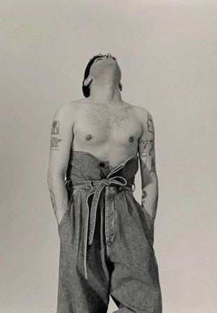 Monochrome portrait of a topless adult male with tattoos, wearing denim overalls, looking upwards in a photo studio with white backdrop.