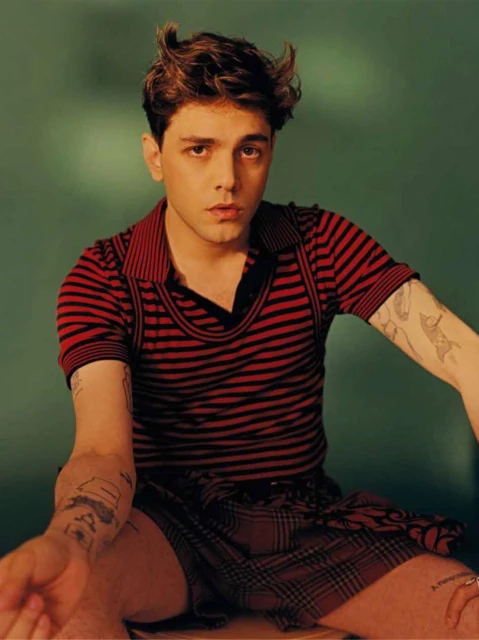 Portrait of a young adult male with tousled brown hair, red and black striped shirt, and tattoos, posing in a studio with green backdrop.