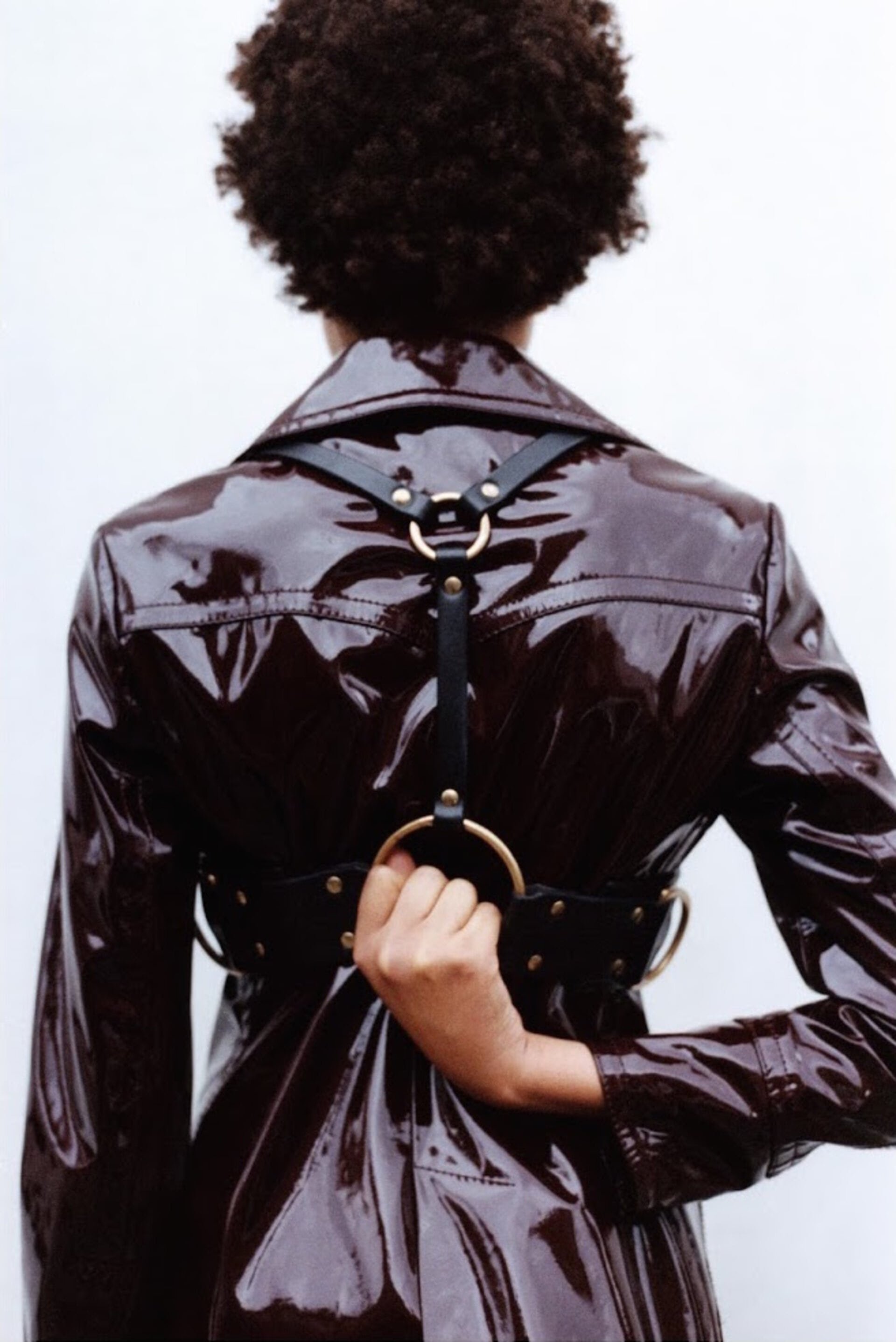 Rear view of a woman with dark curly hair wearing a shiny dark brown coat and a black leather harness with gold rings against a white background.