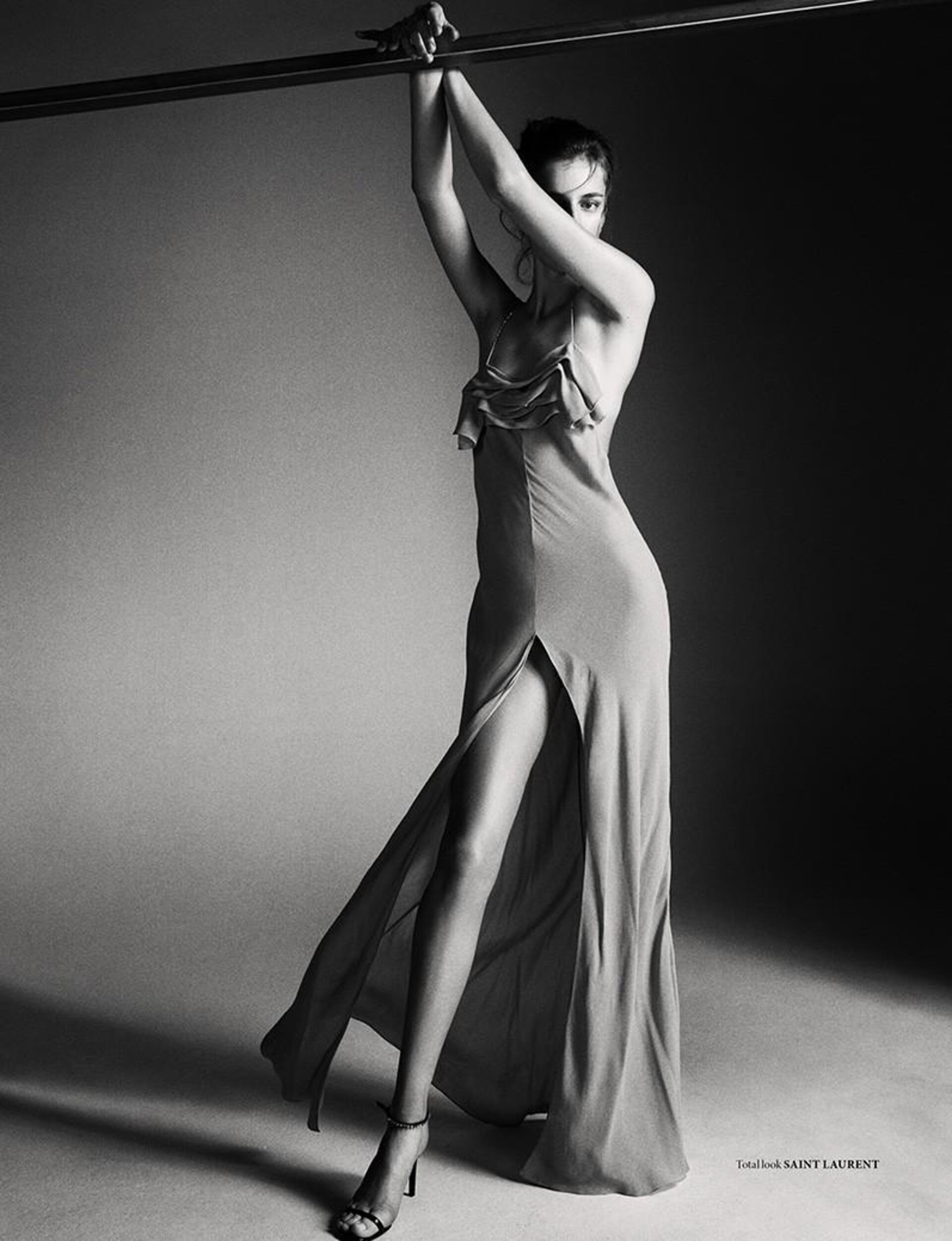 Model in a long slip dress with a thigh-high slit holding onto an overhead bar in a monochrome studio setting styled by Lorenzo Posocco.