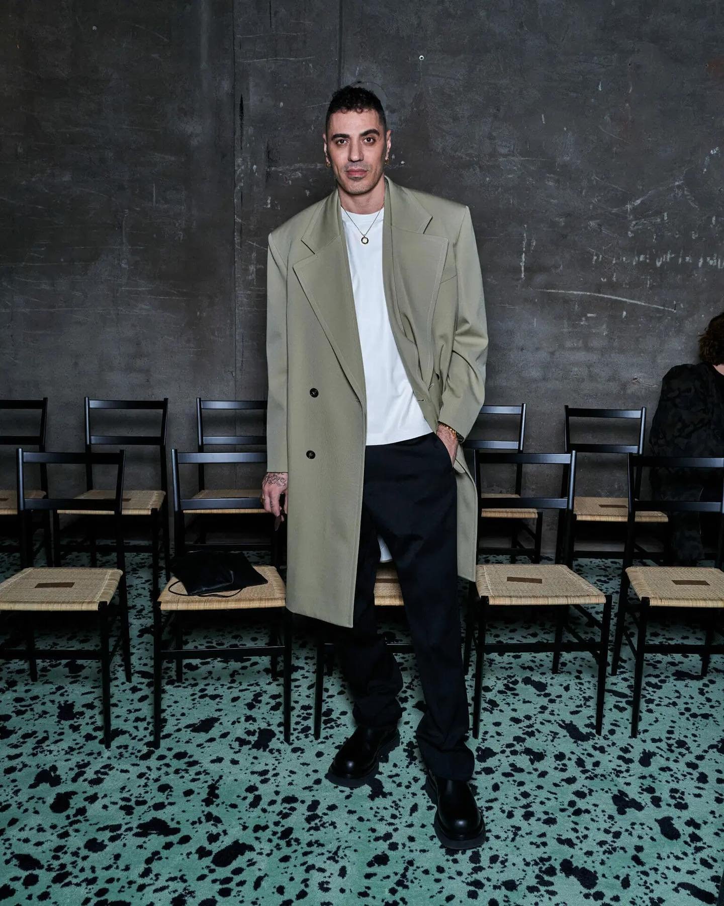 Full-length portrait of a slender adult male in a khaki trench coat, black pants, and black shoes, seated chairs, and a textured wall.
