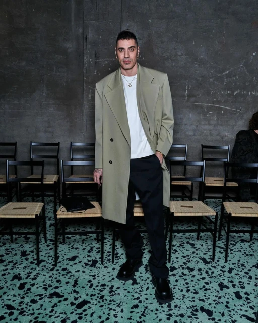 Full-length portrait of a slender adult male in a khaki trench coat, black pants, and black shoes, seated chairs, and a textured wall.