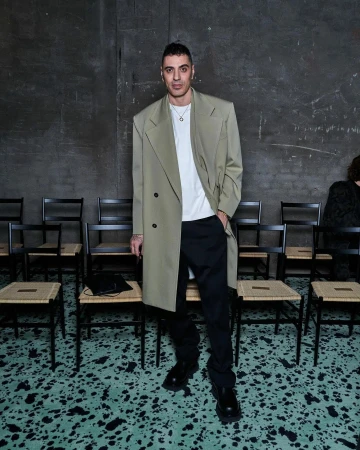 Full-length portrait of a slender adult male in a khaki trench coat, black pants, and black shoes, seated chairs, and a textured wall.