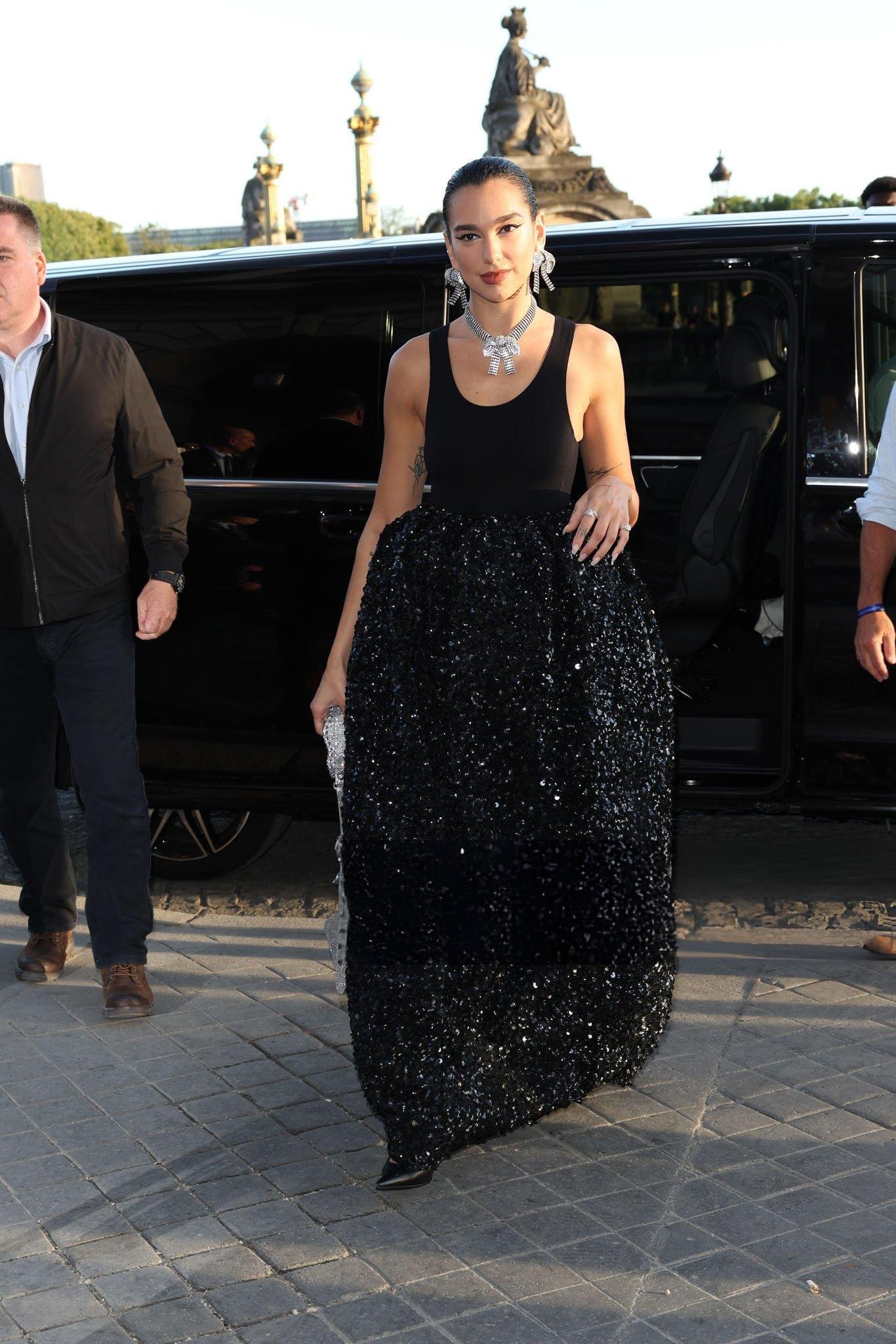 Woman in black gown exits car in Paris with security. The background includes architectural elements and paving stones.