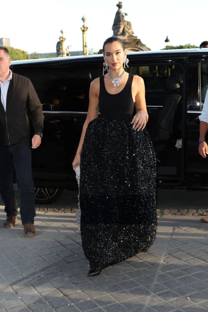 Woman in black gown exits car in Paris with security. The background includes architectural elements and paving stones.