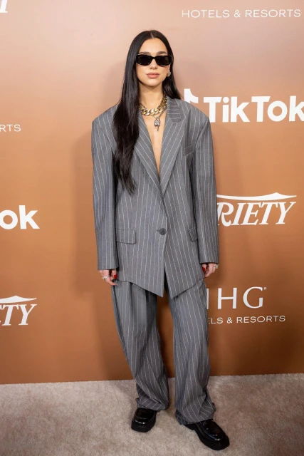Dua Lipa poses in an oversized pinstripe suit with black shoes, sunglasses, and a gold chain necklace, standing against a gradient background.
