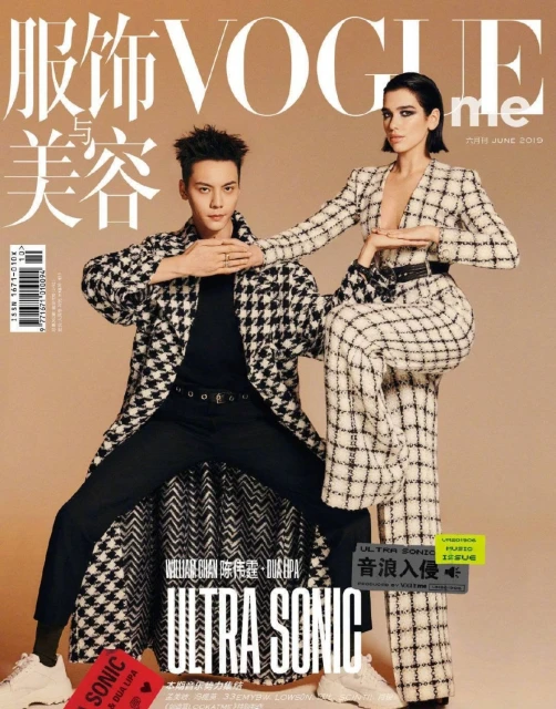 Vogue cover featuring a stylish adult male in a patterned coat with a fashionable adult female in a checkered suit posing indoors.